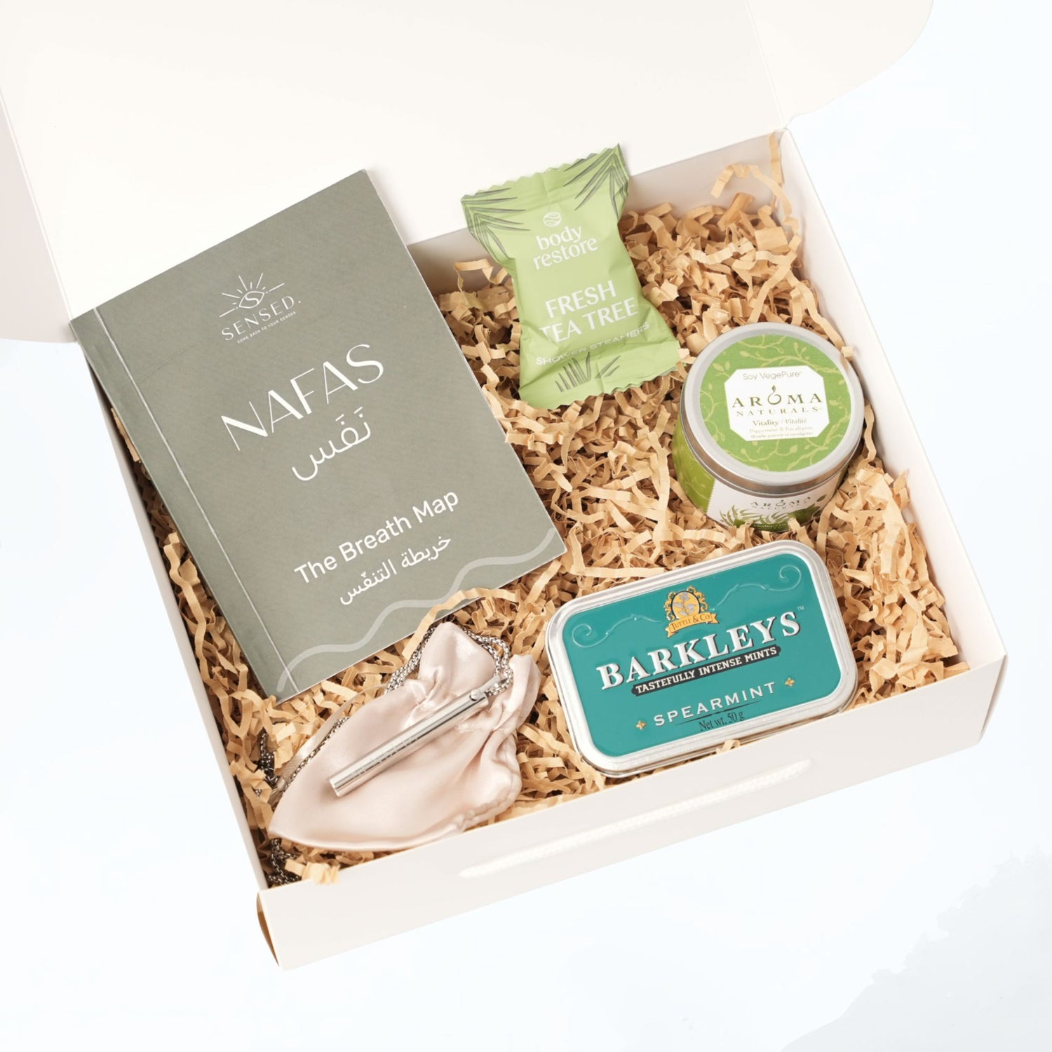 Open Nafas gift box containing the Breath Map booklet, tea tree shower steamer, soy candle, spearmint mints, and Exhale Pendant on shredded kraft paper.