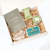 Open Nafas gift box containing the Breath Map booklet, tea tree shower steamer, soy candle, spearmint mints, and Exhale Pendant on shredded kraft paper.